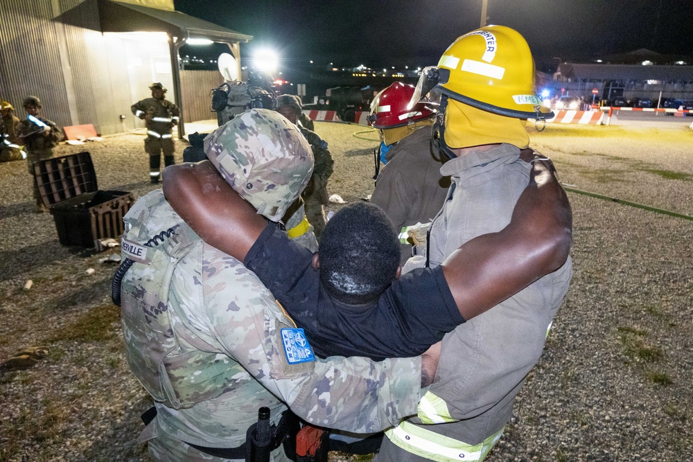 A nighttime emergency response exercise or incident showing military personnel and firefighters assisting an individual. A soldier in camouflage uniform with an MP (Military Police) patch and a firefighter in yellow helmet are supporting a person between them. The scene appears to take place on a military installation based on the uniforms, equipment, and infrastructure visible.