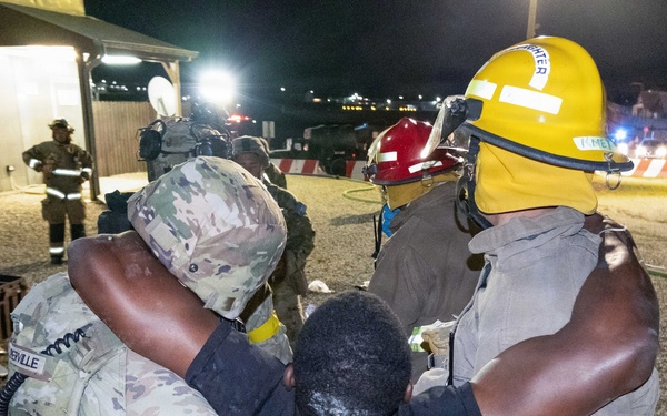 A nighttime emergency response exercise or incident showing military personnel and firefighters assisting an individual. A soldier in camouflage uniform with an MP (Military Police) patch and a firefighter in yellow helmet are supporting a person between them. The scene appears to take place on a military installation based on the uniforms, equipment, and infrastructure visible.