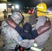 A nighttime emergency response exercise or incident showing military personnel and firefighters assisting an individual. A soldier in camouflage uniform with an MP (Military Police) patch and a firefighter in yellow helmet are supporting a person between them. The scene appears to take place on a military installation based on the uniforms, equipment, and infrastructure visible.