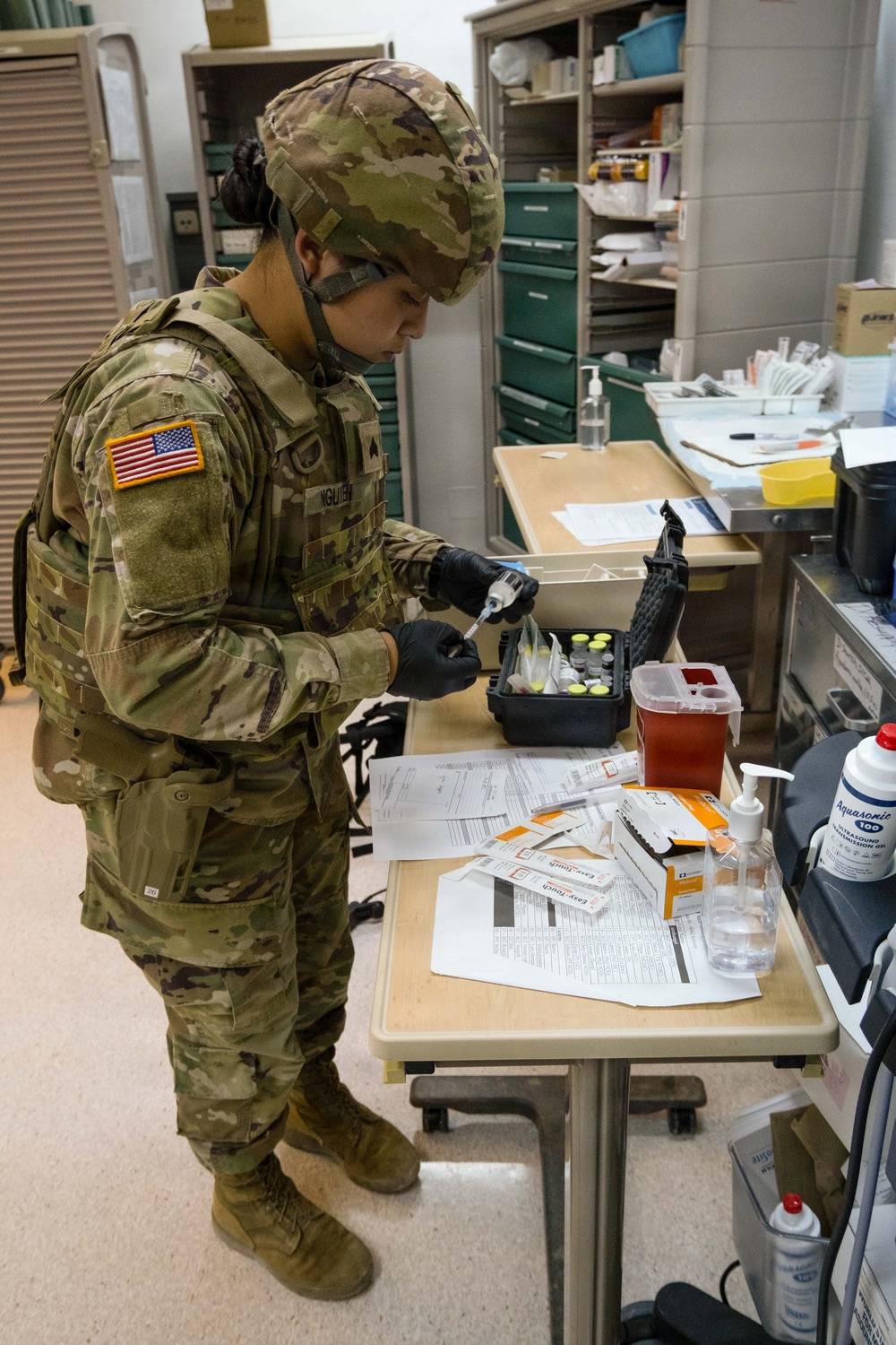 A U.S. Army soldier wearing OCP (Operational Camouflage Pattern) uniform with the name tape 'NGUYEN' is working at a medical supply cart in what appears to be a military medical facility. The soldier is wearing black nitrile gloves and a face mask while handling syringes and vials from a medical kit case. The workspace contains various medical supplies, paperwork, and equipment consistent with a clinical or field medical setting.
