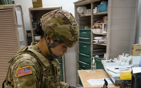 A U.S. Army soldier wearing OCP (Operational Camouflage Pattern) uniform with the name tape 'NGUYEN' is working at a medical supply cart in what appears to be a military medical facility. The soldier is wearing black nitrile gloves and a face mask while handling syringes and vials from a medical kit case. The workspace contains various medical supplies, paperwork, and equipment consistent with a clinical or field medical setting.