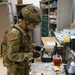 A U.S. Army soldier wearing OCP (Operational Camouflage Pattern) uniform with the name tape 'NGUYEN' is working at a medical supply cart in what appears to be a military medical facility. The soldier is wearing black nitrile gloves and a face mask while handling syringes and vials from a medical kit case. The workspace contains various medical supplies, paperwork, and equipment consistent with a clinical or field medical setting.