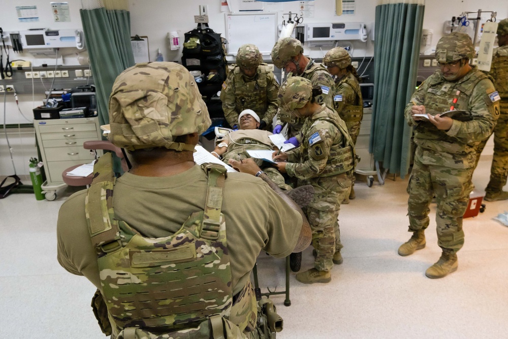 Military personnel in OCP (Operational Camouflage Pattern) uniforms are conducting what appears to be a medical training exercise in a clinical or field hospital setting. A simulated casualty is lying on a gurney with a head bandage while multiple soldiers attend to them, taking notes and performing assessments. The setting includes medical equipment, supply carts, and curtained treatment bays consistent with a military medical facility.