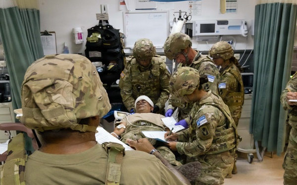 Military personnel in OCP (Operational Camouflage Pattern) uniforms are conducting what appears to be a medical training exercise in a clinical or field hospital setting. A simulated casualty is lying on a gurney with a head bandage while multiple soldiers attend to them, taking notes and performing assessments. The setting includes medical equipment, supply carts, and curtained treatment bays consistent with a military medical facility.
