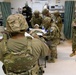 Military personnel in OCP (Operational Camouflage Pattern) uniforms are conducting what appears to be a medical training exercise in a clinical or field hospital setting. A simulated casualty is lying on a gurney with a head bandage while multiple soldiers attend to them, taking notes and performing assessments. The setting includes medical equipment, supply carts, and curtained treatment bays consistent with a military medical facility.