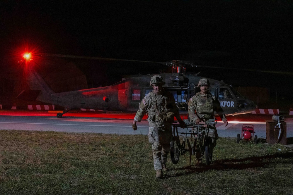 Two U.S. Army soldiers in full combat gear are carrying a stretcher/litter away from a UH-60 Black Hawk helicopter at night. The helicopter bears the marking 'KFOR' (Kosovo Force), indicating this is a NATO-led peacekeeping operation in Kosovo. The scene is illuminated by a red navigation/warning light on the aircraft and ambient ground lighting.