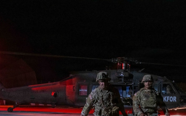 Two U.S. Army soldiers in full combat gear are carrying a stretcher/litter away from a UH-60 Black Hawk helicopter at night. The helicopter bears the marking 'KFOR' (Kosovo Force), indicating this is a NATO-led peacekeeping operation in Kosovo. The scene is illuminated by a red navigation/warning light on the aircraft and ambient ground lighting.