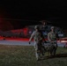 Two U.S. Army soldiers in full combat gear are carrying a stretcher/litter away from a UH-60 Black Hawk helicopter at night. The helicopter bears the marking 'KFOR' (Kosovo Force), indicating this is a NATO-led peacekeeping operation in Kosovo. The scene is illuminated by a red navigation/warning light on the aircraft and ambient ground lighting.
