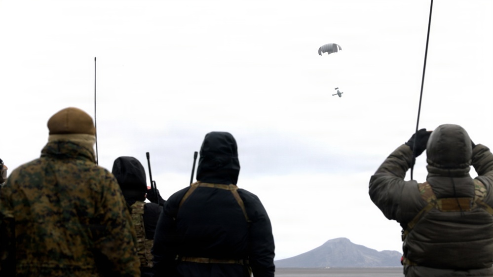 A group of military personnel in cold-weather gear observe an aerial object descending under a parachute in an overcast sky. The scene appears to be a parachute drop or airdrop exercise conducted in a flat, arid terrain with a mountain visible in the background. Personnel are holding radio antennas and communication equipment, suggesting they are coordinating or monitoring the drop.