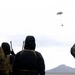 A group of military personnel in cold-weather gear observe an aerial object descending under a parachute in an overcast sky. The scene appears to be a parachute drop or airdrop exercise conducted in a flat, arid terrain with a mountain visible in the background. Personnel are holding radio antennas and communication equipment, suggesting they are coordinating or monitoring the drop.