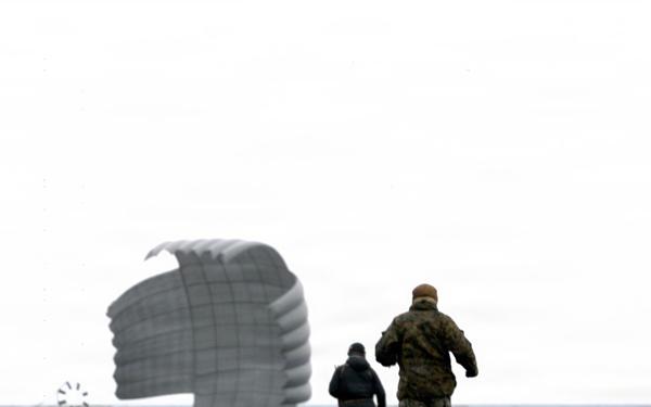 Two individuals are running across a dark, flat terrain toward a deployed parachute and what appears to be a landed cargo or equipment container. One person is wearing military camouflage uniform while the other is in darker civilian-style clothing. The scene appears to be a military airdrop recovery operation conducted under overcast sky conditions.