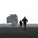 Two individuals are running across a dark, flat terrain toward a deployed parachute and what appears to be a landed cargo or equipment container. One person is wearing military camouflage uniform while the other is in darker civilian-style clothing. The scene appears to be a military airdrop recovery operation conducted under overcast sky conditions.