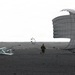 Two individuals are on a dark, flat terrain attempting to manage a large deployed parachute that is being inflated or dragged by wind. A piece of metal equipment or structure lies toppled on the ground to the left, and a smaller parachute or drogue chute is visible in the background. The overcast sky and dark ground suggest a remote, possibly coastal or volcanic landscape.