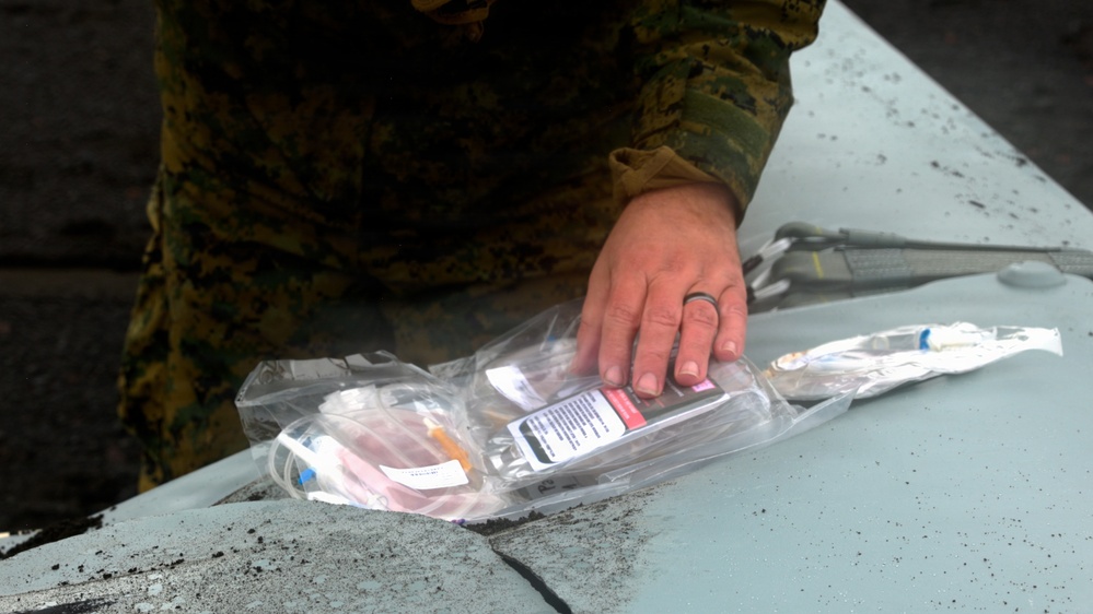 A person wearing military camouflage uniform is handling a clear plastic medical supply bag on a light blue surface. The bag contains what appears to be syringes, tubing, and a labeled medical vial or package. The individual is wearing a ring on their finger and appears to be organizing or retrieving medical supplies.