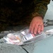 A person wearing military camouflage uniform is handling a clear plastic medical supply bag on a light blue surface. The bag contains what appears to be syringes, tubing, and a labeled medical vial or package. The individual is wearing a ring on their finger and appears to be organizing or retrieving medical supplies.