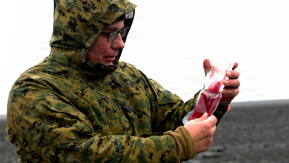 A person wearing digital camouflage military uniform and glasses is holding a clear plastic bag containing a red liquid substance. The individual has their hood up, suggesting outdoor conditions in cold or wet weather. The background appears to be an overcast, open terrain environment.