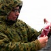 A person wearing digital camouflage military uniform and glasses is holding a clear plastic bag containing a red liquid substance. The individual has their hood up, suggesting outdoor conditions in cold or wet weather. The background appears to be an overcast, open terrain environment.