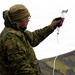 A military personnel wearing MARPAT digital camouflage uniform holds up a blood bag (IV blood product) in an outdoor, mountainous environment. The setting appears cold and overcast, consistent with a northern or arctic training environment. This appears to be a field medical training exercise or demonstration involving blood product handling.