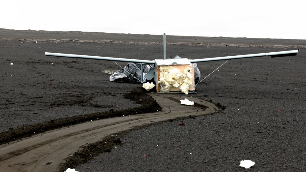 A small aircraft wreckage is resting on a dark volcanic or black sand terrain, appearing to have crash-landed or been severely damaged. The fuselage is torn open, exposing white foam insulation material, while the wings remain largely intact. Tire or skid tracks in the dark sand suggest the aircraft slid along the surface before coming to rest.