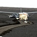 A small aircraft wreckage is resting on a dark volcanic or black sand terrain, appearing to have crash-landed or been severely damaged. The fuselage is torn open, exposing white foam insulation material, while the wings remain largely intact. Tire or skid tracks in the dark sand suggest the aircraft slid along the surface before coming to rest.
