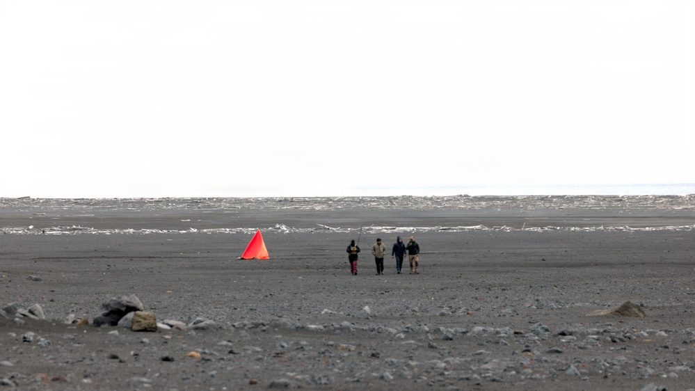 Four people are walking across a vast, dark sandy or volcanic terrain toward a distant shoreline or frozen body of water. A bright orange triangular marker or cone is visible to their left, likely serving as a navigation or survey marker. The landscape is barren and flat, with overcast skies and scattered rocks in the foreground.