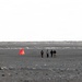 Four people are walking across a vast, dark sandy or volcanic terrain toward a distant shoreline or frozen body of water. A bright orange triangular marker or cone is visible to their left, likely serving as a navigation or survey marker. The landscape is barren and flat, with overcast skies and scattered rocks in the foreground.