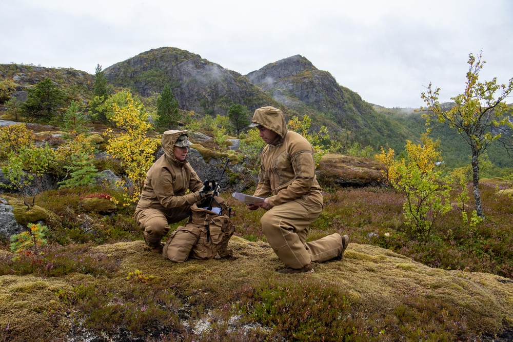 Two military personnel in tan/coyote-colored uniforms are kneeling on a moss-covered rocky outcrop in a mountainous, subarctic or Nordic landscape. One individual appears to be operating a radio communications device while the other reviews a document or map. The autumn foliage, rocky peaks, low cloud cover, and tundra-like vegetation suggest a Scandinavian or northern European training environment.