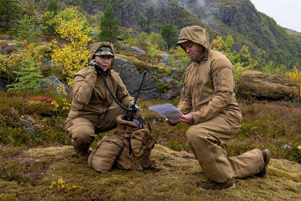 Two military personnel in tan/coyote-brown uniforms are positioned on a moss-covered rocky terrain in an autumn mountainous landscape. One individual is using a radio handset while the other reviews what appears to be a document or map in a clear protective sleeve. The setting suggests a field exercise or operational activity in a Nordic or Scandinavian-type environment.
