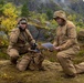 Two military personnel in tan/coyote-brown uniforms are positioned on a moss-covered rocky terrain in an autumn mountainous landscape. One individual is using a radio handset while the other reviews what appears to be a document or map in a clear protective sleeve. The setting suggests a field exercise or operational activity in a Nordic or Scandinavian-type environment.