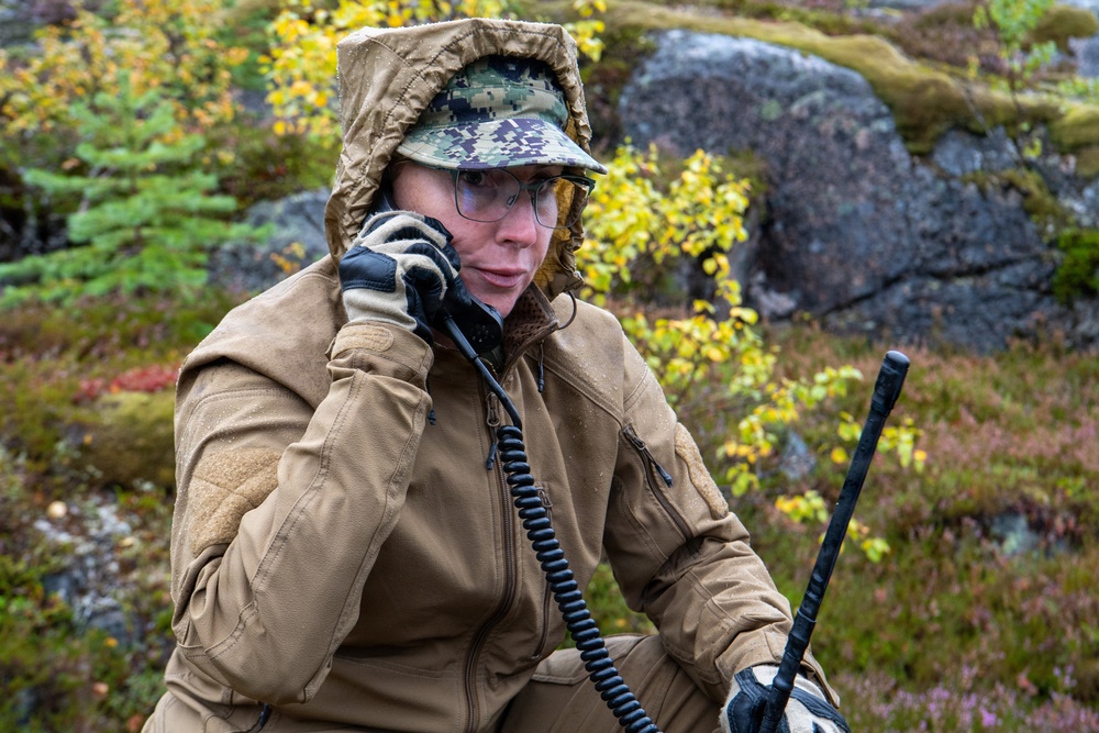 A person in military or tactical gear is using a handheld radio communication device in an outdoor wilderness setting. They are wearing a tan/coyote brown jacket with a digital camouflage cap and protective eyewear. The environment appears to be a rocky, autumnal Nordic or subarctic landscape.