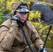 A person in military or tactical gear is using a handheld radio communication device in an outdoor wilderness setting. They are wearing a tan/coyote brown jacket with a digital camouflage cap and protective eyewear. The environment appears to be a rocky, autumnal Nordic or subarctic landscape.