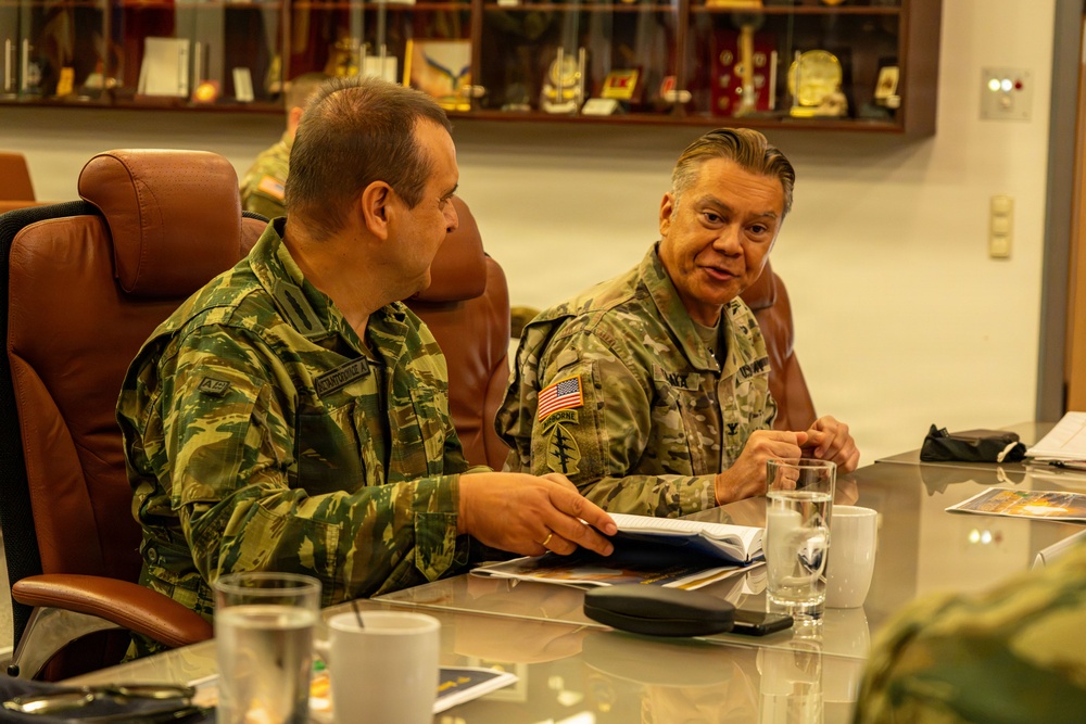 Two military officers in camouflage uniforms are seated at a conference table engaged in discussion. The officer on the right wears a U.S. Army uniform with an American flag patch and Airborne/Special Forces insignia, while the officer on the left wears a different camouflage pattern suggesting a foreign military. The setting appears to be a formal military meeting room with a display case of memorabilia and awards visible in the background.