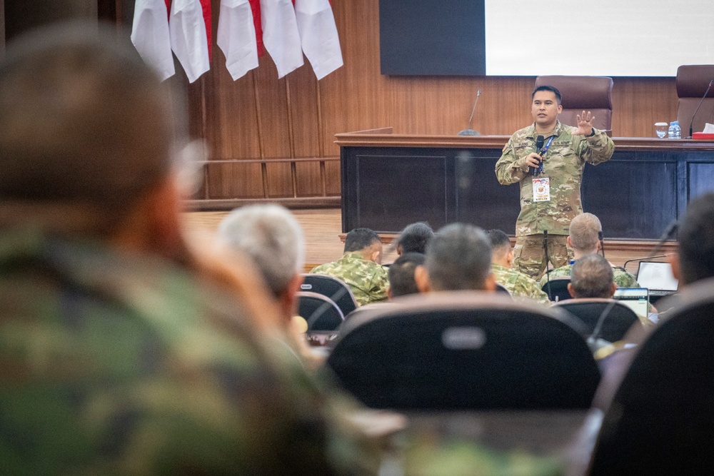 A U.S. Army soldier is delivering a briefing or presentation to an audience of military personnel in a formal conference or auditorium setting. The speaker is standing at the front of the room, gesturing with his hands while addressing seated attendees in military uniforms. Red and white flags are visible in the background, suggesting a multinational or joint military exercise environment.