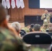 A U.S. Army soldier is delivering a briefing or presentation to an audience of military personnel in a formal conference or auditorium setting. The speaker is standing at the front of the room, gesturing with his hands while addressing seated attendees in military uniforms. Red and white flags are visible in the background, suggesting a multinational or joint military exercise environment.