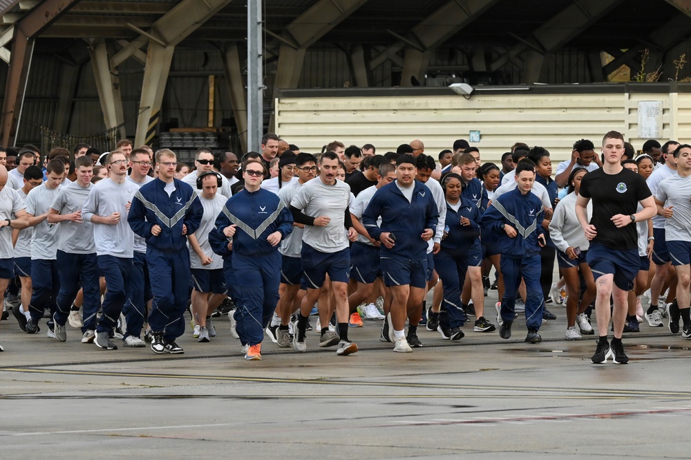 A large group of military personnel, appearing to be U.S. Air Force members based on visible insignia and uniforms, are participating in a group run or physical training event. Participants are wearing a mix of blue Air Force physical training (PT) uniforms and gray t-shirts with blue shorts. The event is taking place on a tarmac or flight line area in front of a large aircraft hangar.