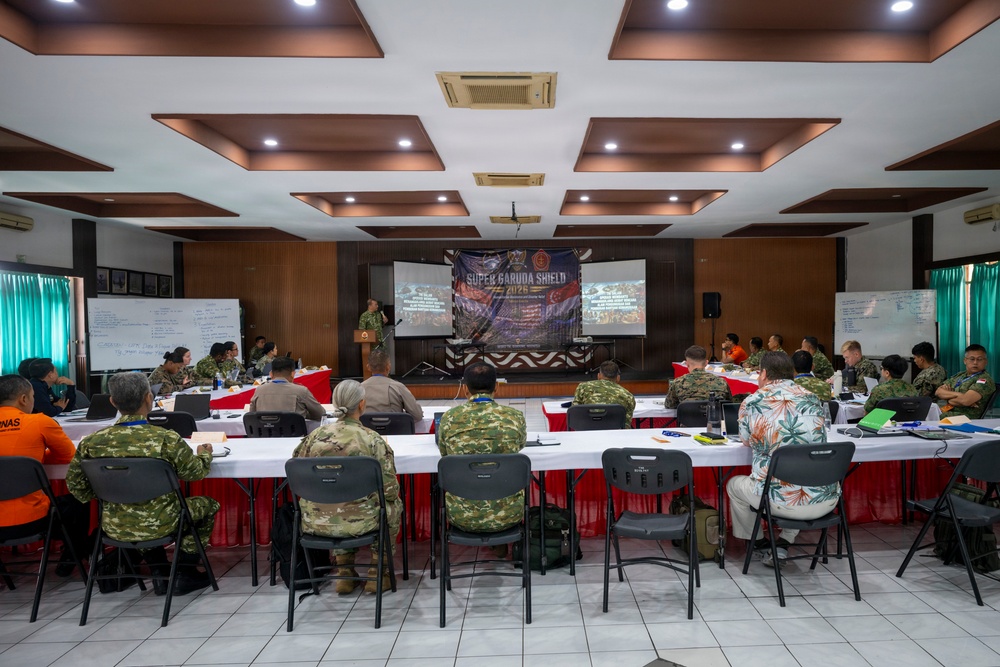 A multinational military briefing or conference is taking place in a formal meeting room. Participants in various military uniforms and civilian attire are seated at U-shaped tables facing a presenter at a podium. A banner reading 'Super Garuda Shield 2025' is prominently displayed at the front of the room.