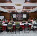 A multinational military briefing or conference is taking place in a formal meeting room. Participants in various military uniforms and civilian attire are seated at U-shaped tables facing a presenter at a podium. A banner reading 'Super Garuda Shield 2025' is prominently displayed at the front of the room.