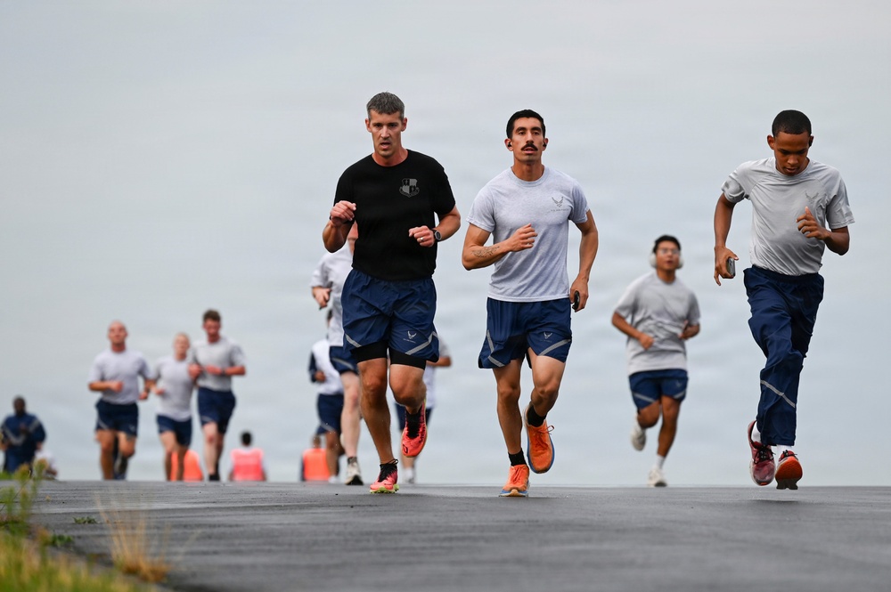 A large group of individuals, appearing to be military personnel based on their uniforms and insignia, are participating in a group run on what appears to be a paved runway or road. Three runners are prominently in the foreground, with one wearing a black t-shirt and two wearing gray shirts with what appear to be U.S. Air Force logos. Orange traffic cones are visible in the background, suggesting an organized physical training event.