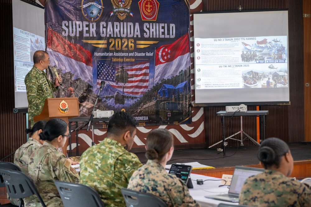 A military briefing or tabletop exercise is taking place under the 'Super Garuda Shield 2026' banner, focused on Humanitarian Assistance and Disaster Relief (HADR). A uniformed officer stands at a podium presenting to a group of multinational military personnel seated at tables. The event appears to be a joint military exercise involving at least the United States, Indonesia, and Singapore, held in Indonesia in 2026.