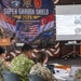 A military briefing or tabletop exercise is taking place under the 'Super Garuda Shield 2026' banner, focused on Humanitarian Assistance and Disaster Relief (HADR). A uniformed officer stands at a podium presenting to a group of multinational military personnel seated at tables. The event appears to be a joint military exercise involving at least the United States, Indonesia, and Singapore, held in Indonesia in 2026.