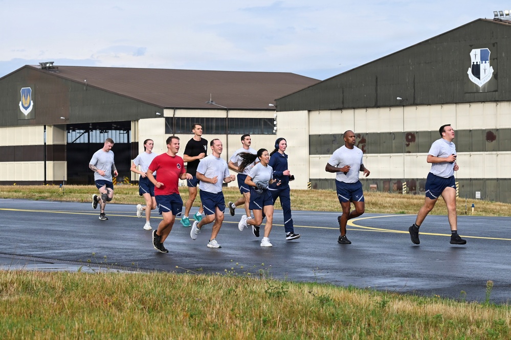 A group of approximately 12 military personnel are running on a paved surface at what appears to be a U.S. Air Force base. The runners are wearing standard Air Force physical training (PT) gear consisting of gray shirts and navy blue shorts, with one individual in a red shirt. Two large aircraft hangars bearing U.S. Air Force shield insignia are visible in the background.
