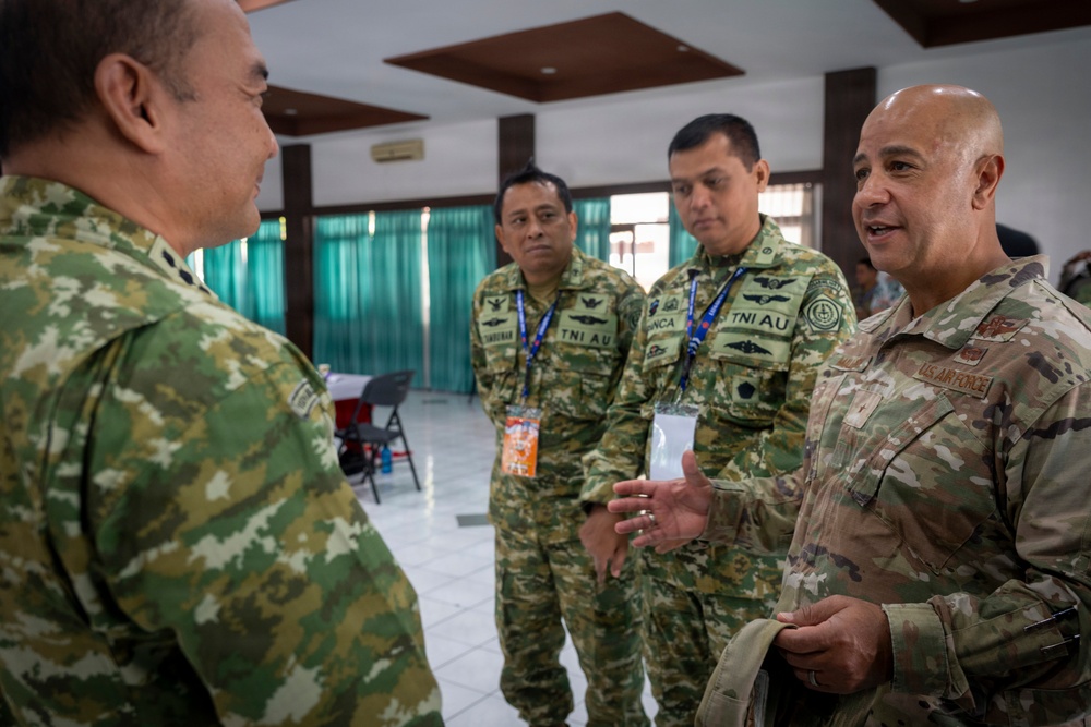 A U.S. Air Force officer in OCP (Operational Camouflage Pattern) uniform is engaged in conversation with several military personnel wearing TNI AU (Indonesian Air Force) uniforms. The setting appears to be an indoor meeting or conference facility, suggesting a bilateral or multilateral military engagement. The interaction appears cordial and professional in nature.