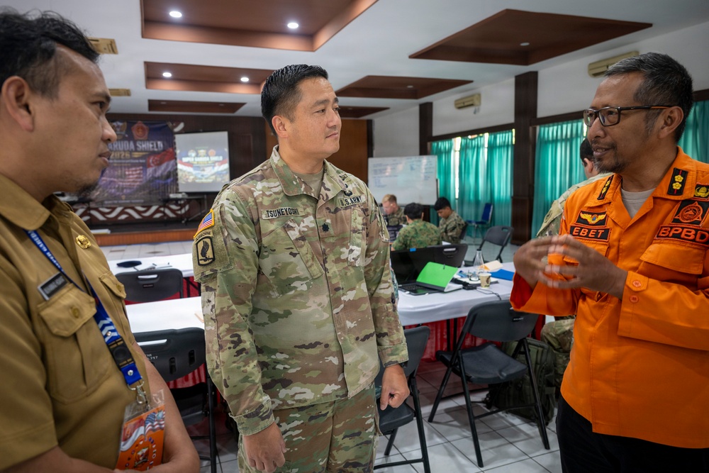 A U.S. Army soldier identified by name tape as 'TSUNEYOSHI' is engaged in conversation with two Indonesian officials in what appears to be a military coordination or briefing room. The setting suggests a joint military exercise or bilateral meeting, with a banner in the background referencing 'GARUDA SHIELD 2025.' The interaction appears cooperative and informal, taking place during a working session.