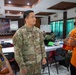 A U.S. Army soldier identified by name tape as 'TSUNEYOSHI' is engaged in conversation with two Indonesian officials in what appears to be a military coordination or briefing room. The setting suggests a joint military exercise or bilateral meeting, with a banner in the background referencing 'GARUDA SHIELD 2025.' The interaction appears cooperative and informal, taking place during a working session.