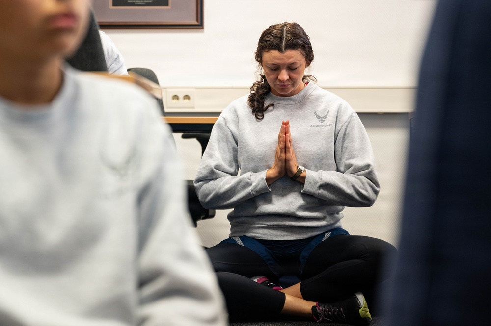 A woman wearing a U.S. Air Force sweatshirt sits cross-legged on the floor in a prayer or namaste hand position with eyes closed, appearing to meditate. She is in what appears to be an indoor room, surrounded by other partially visible individuals also seated. The setting suggests a group mindfulness or yoga session.