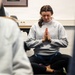A woman wearing a U.S. Air Force sweatshirt sits cross-legged on the floor in a prayer or namaste hand position with eyes closed, appearing to meditate. She is in what appears to be an indoor room, surrounded by other partially visible individuals also seated. The setting suggests a group mindfulness or yoga session.