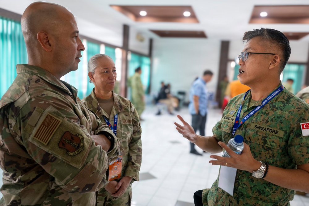 Three military personnel are engaged in conversation in what appears to be a conference or meeting facility. Two individuals wearing US military multicam uniforms are speaking with a third person in Singapore Armed Forces digital camouflage uniform. The setting suggests a multinational military exercise or conference event, as indicated by lanyards and name badges worn by the participants.