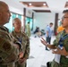 Three military personnel are engaged in conversation in what appears to be a conference or meeting facility. Two individuals wearing US military multicam uniforms are speaking with a third person in Singapore Armed Forces digital camouflage uniform. The setting suggests a multinational military exercise or conference event, as indicated by lanyards and name badges worn by the participants.