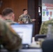 A military personnel in camouflage uniform is delivering a briefing or presentation at a podium to an audience of other military members. The projected slide behind the speaker displays an organizational chart titled 'Organization' with references to what appears to be the Hawaii National Guard structure. The setting appears to be a formal military conference or training room.
