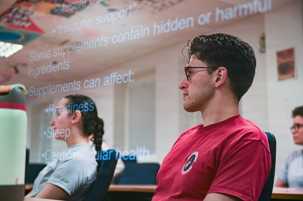 A group of students or attendees are seated in a classroom or lecture setting, attentively watching a presentation. A projected slide is visible overhead discussing dietary supplements, their potential risks, and their effects on health. The foreground subject is a young male wearing glasses and a red t-shirt.